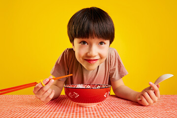 Cheerful young boy using chopsticks and spoon, sitting at table and eating ramen, noodle soup...