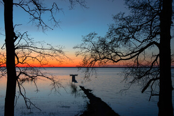 orange sunset on a lake between two trees with a flooded boardwalk on a spring evening and an...