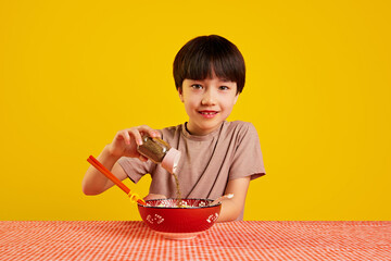 Little boy, child sitting at table, eating ramen soup and adding spices, pepper to his meal against yellow background. Spicy taste. Concept of food, childhood, emotions, meal, menu, pop art