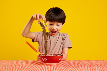 Excited young boy pouring generous amount of pepper into bowl of noodles, ramen soup against yellow background. Japanese spicy dinner. Concept of food, childhood, emotions, meal, menu, pop art