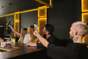 Young elegantly dressed ladies and men pose for selfies at bar. Blonde man with tattoo on hand and neck raises glass with cocktail pointing at phone camera
