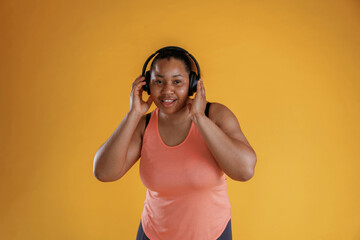 Touching headphones, listening to music. African American woman is in the studio against yellow background