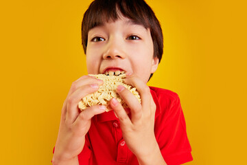 Young boy, child in a red polo shirt enjoying noodles, eating with his hands against yellow background. Concept of food, childhood, emotions, meal, menu, pop art. Close-up portrait