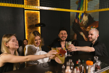 Joyful friends sip alcoholic cocktails and raise glasses to clink in contemporary pub. Young people...