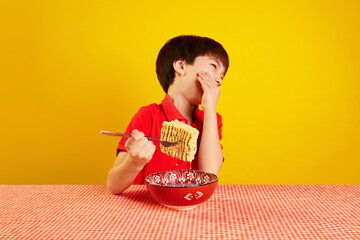 Cheerful child in red polo shirt sitting at table, laughing and eating instant noodles with fork against yellow background. Concept of food, childhood, emotions, meal, menu, pop art