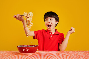 Cheerful emotional child playfully lifting noodles with fork against yellow background. Playful eating. Fast dinner ideas. Concept of food, childhood, emotions, meal, menu, pop art
