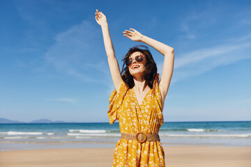 Joyful woman in a sunny yellow dress celebrating on beautiful sandy beach with arms raised high in...