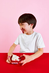 Smiling little Korean boy sitting with dirty face in cake cream, eating with pleasure red velvet cake against pink background. Concept of food, childhood, emotions, meal, menu, pop art