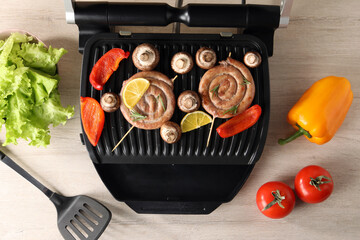 Electric grill with homemade sausages, vegetables and spatula on wooden table, flat lay