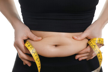 Woman with measuring tape touching belly fat on white background, closeup. Overweight problem