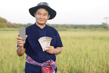 Asian man farmer is at paddy field, wears hat, holds Thai banknote money and smart phone. Concept,...