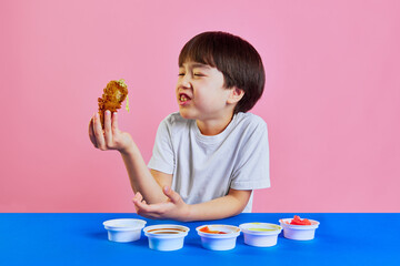 Little boy, Korean kid sitting at table and emotionally eating fried chicken with different sauces against pink background. Concept of food, childhood, emotions, meal, menu, pop art