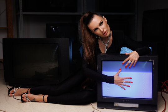 A beautiful girl in a black jumpsuit with an open neckline and high heels, against the backdrop of old working TVs standing in a row. A portrait of girl with bright makeup, winged eyes, big lips.