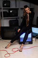 A beautiful girl in a black jumpsuit with an open neckline and high heels, against the backdrop of old working TVs standing in a row. A portrait of girl with bright makeup, winged eyes, big lips.