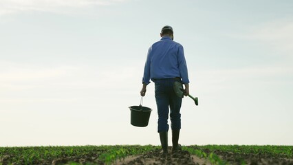 Farmer with shovel bucket walks through agricultural field. Tired exhausted man male businessman...
