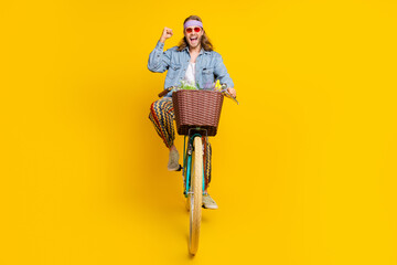 Full size photo of cool young man ride bike raise fist empty space wear denim shirt isolated on yellow color background