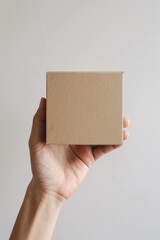 Hand holding a square brown paper box in front of beige background