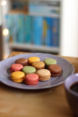 Purple plate filled with pastel macarons, cup of tea or coffee, vintage books and reading glasses on the table. Colorful bookcase in the background. Selective focus.