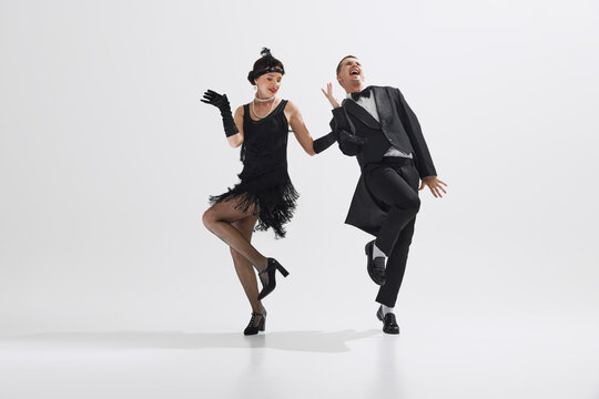 Woman in black flapper dress dancing with man in classic black suit, showcasing lively spirit of the 1920s isolated over white studio background. Concept of art, retro and vintage, entertainment, 20s