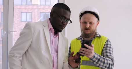 Diverse duo, consisting of homeowner and construction manager, engage deeply over smartphone...