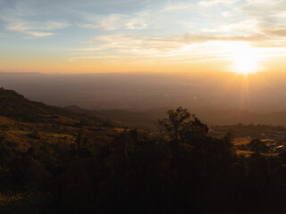 Obraz premium The sun rises, casting a golden light over a mountainous landscape with silhouetted trees, creating a stunning morning scene.