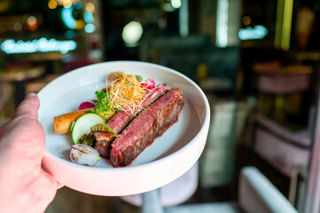 A hand holds a plate of delicious steak, garnished with fresh vegetables and herbs, in a cozy,...