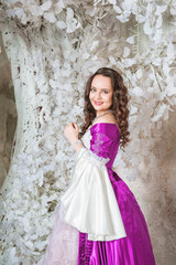 Beautiful smiling woman in fantasy white and purple rococo style medieval dress standing near tree with white flowers