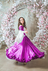 Fototapeta premium Happy beautiful woman in fantasy white and purple rococo style medieval dress dancing near decoration with white flowers