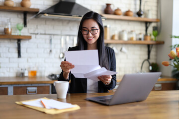 An Asian businesswoman happily reads a finance letter at her desk, managing paperwork in her office.