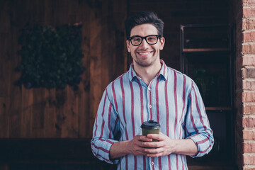 Portrait of nice young man toothy smile hold coffee wear striped shirt loft interior business center indoors