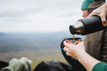 Person pouring tea from a thermos, making a travel gesture under the sky