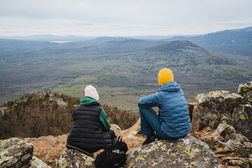 Two people sitting on bedrock atop mountain, enjoying sky and landscape