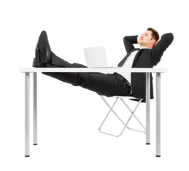 A man in a suit relaxing with his feet on a desk, next to a laptop, against a white background, depicting leisure at work