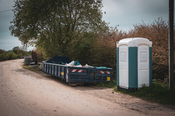 Next to a road is a large blue container for construction waste and a construction site toilet