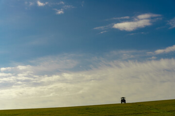 A tractor is sitting on a field of grass. The sky is clear and blue, with a few clouds scattered throughout. The scene is peaceful and serene