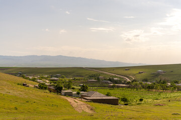 A rural landscape with a road running through it. The road is lined with houses and a few trees. The sky is mostly clear, with a few clouds scattered throughout