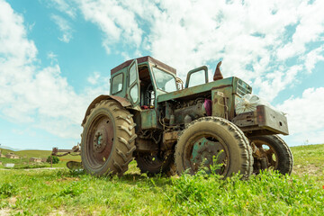 An old green tractor is parked in a field. The tractor is rusty and has a worn-out appearance. The sky is cloudy, and the grass is lush and green