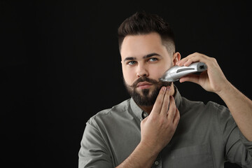 Handsome young man trimming beard on black background. Space for text