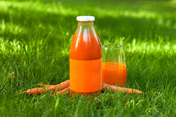 a glass bottle of carrot juice with a glass stands on the lawn in the grass.