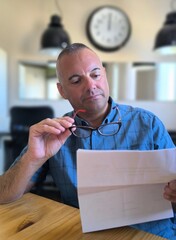 Portrait of a young adult man sitting at the table with eyeglasses in one hand and papers in the...