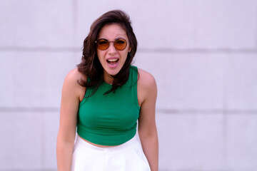 Beautiful girl in a green top, white shorts and sunglasses posing. Aggressively yells while looking at the camera. Against the background of a gray wall
