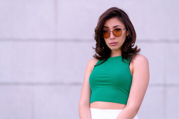 Beautiful girl in a green top, white shorts and sunglasses posing. Looks at the camera in disbelief. Against the background of a gray wall