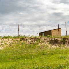 A fence surrounds a small building with a door. The building is made of stone and is located on a hill. The sky is cloudy and the grass is green