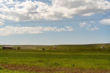 Fototapeta premium A large, open field with a few houses in the distance. The sky is mostly clear, with a few clouds scattered throughout. Scene is peaceful and serene, with the vast expanse of grass