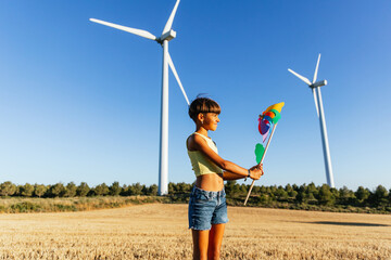 Girl standing alone in field with pinwheel shining in sunbeam. - obrazy, fototapety, plakaty