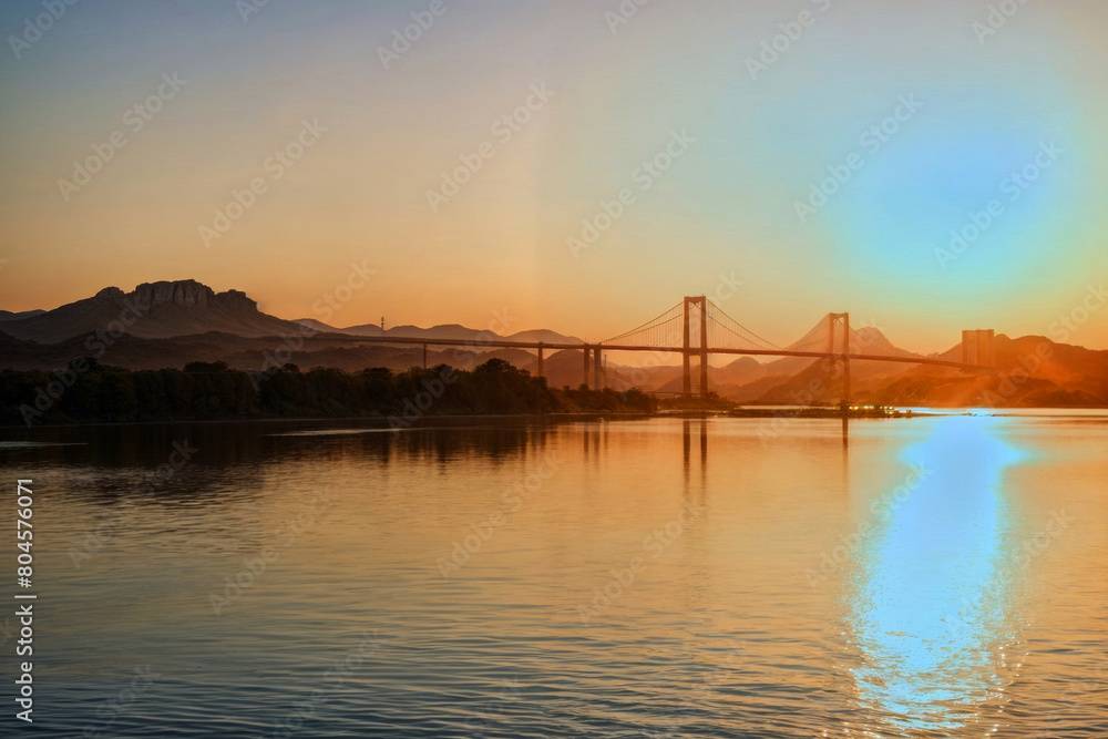 Wall mural A bridge spans a river with a beautiful sunset in the background. The bridge is illuminated by the sun, creating a warm and serene atmosphere