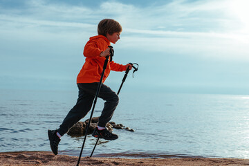 Happy boy hiker jumps with trekking poles on lake shore