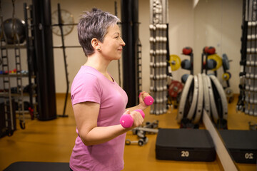 Side view of adult smiling woman doing sport exercise with dumbbells for biceps