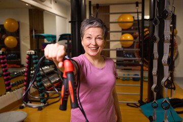 Mature smiling woman holding skipping rope and looking at camera in gym