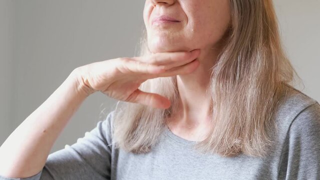 A woman with gray hair massages and pats her neck with sagging skin with her hands. Concept of loss of collagen and sagging skin in adulthood.